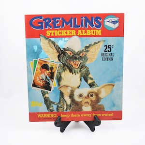 May include: A vintage Gremlins sticker album with a red and blue cover. The cover features images of Gremlins characters and the text "Gremlins Sticker Album". The album is displayed on a black stand.