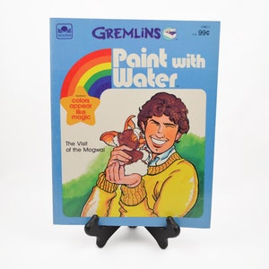 May include: A blue and white book cover with the title "GREMLINS Paint with Water" and a rainbow arc. The cover features a boy holding a small furry creature. The text "colors appear like magic" is in a yellow star.