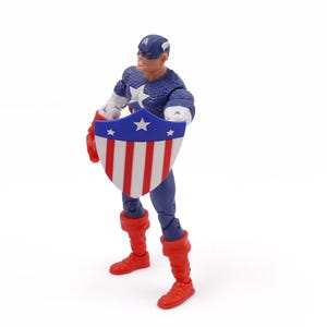 K&ouml;nnte beinhalten: Eine Captain America-Actionfigur, die einen Schild mit roten und wei&szlig;en Streifen und einem blauen Sternenfeld h&auml;lt. Die Figur tr&auml;gt ein blau-wei&szlig;es Kost&uuml;m mit roten Stiefeln und Handschuhen. Der Hintergrund ist wei&szlig;.