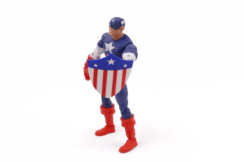 K&ouml;nnte beinhalten: Eine Captain America-Actionfigur, die einen Schild mit roten und wei&szlig;en Streifen und einem blauen Sternenfeld h&auml;lt. Die Figur tr&auml;gt ein blau-wei&szlig;es Kost&uuml;m mit roten Stiefeln und Handschuhen. Der Hintergrund ist wei&szlig;.