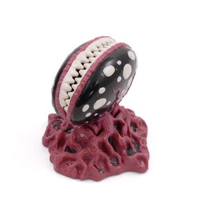 May include: A stylized, three-dimensional sculpture of a carnivorous plant. The plant has a black head with white polka dots and a mouth full of white teeth. The base is a textured, reddish-brown color.