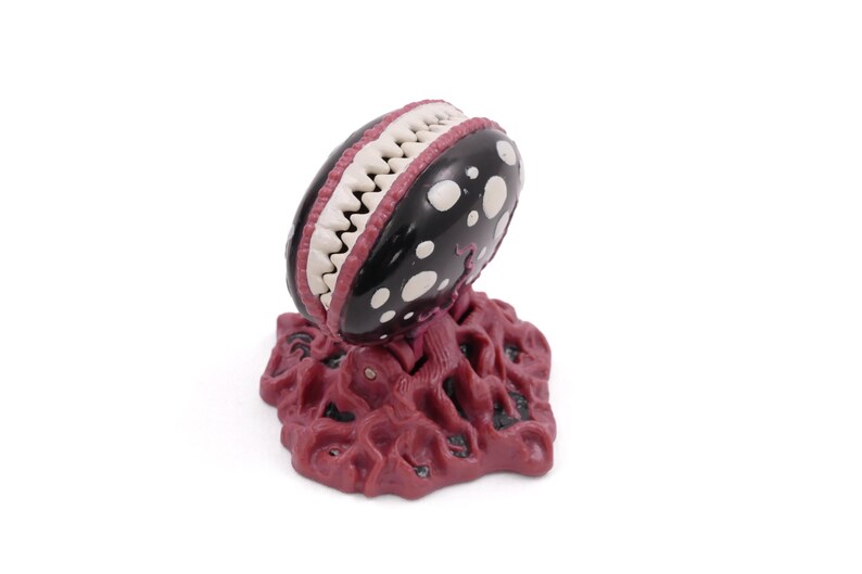 May include: A stylized, three-dimensional sculpture of a carnivorous plant. The plant has a black head with white polka dots and a mouth full of white teeth. The base is a textured, reddish-brown color.