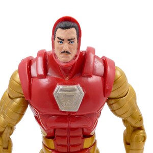 May include: A close-up of an Iron Man action figure. The figure has a red and gold color scheme, with a red helmet and chest piece, and gold arms. The face is visible, with a mustache and blue eyes.