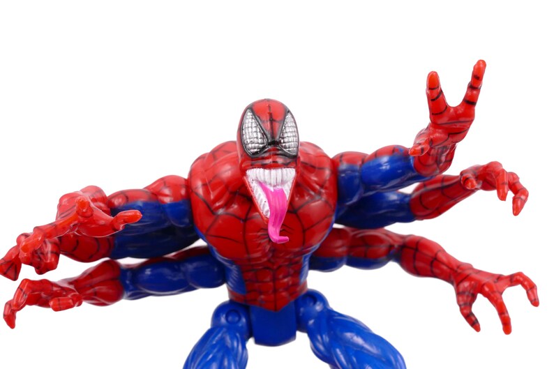 May include: A red, blue, and white action figure of a superhero with multiple arms. The figure has a red and blue costume, a white mouth with a pink tongue, and a black and white mask.