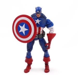 May include: A Captain America action figure stands with a shield and a red gauntlet. The figure is in a blue, white, and red costume, with a star on the chest and shield. The shield is round with a white star in a blue circle.