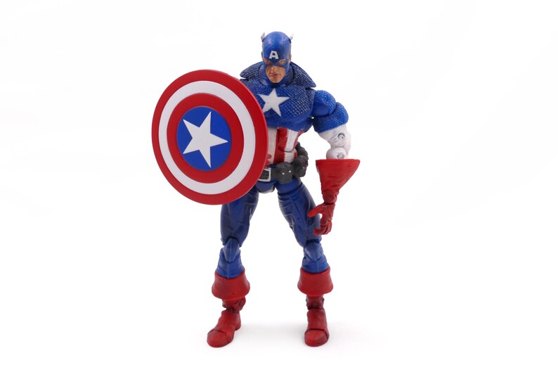 May include: A Captain America action figure stands with a shield and a red gauntlet. The figure is in a blue, white, and red costume, with a star on the chest and shield. The shield is round with a white star in a blue circle.