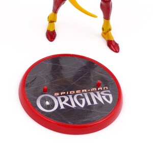 May include: A red and black circular stand with the words "Spider-Man Origins" printed on it. The stand has three small pegs on the top.