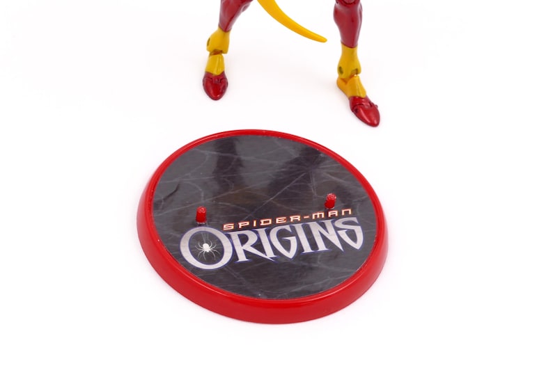 May include: A red and black circular stand with the words "Spider-Man Origins" printed on it. The stand has three small pegs on the top.