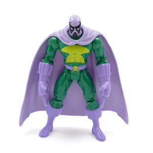 Puede incluir: Una figura de acci&oacute;n verde con un emblema de pecho amarillo, capa, guantes y botas moradas. La figura tiene una m&aacute;scara morada y una cara negra. La figura est&aacute; sobre una superficie blanca.