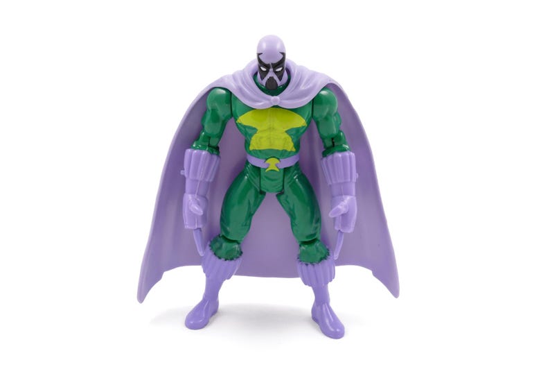 Puede incluir: Una figura de acci&oacute;n verde con un emblema de pecho amarillo, capa, guantes y botas moradas. La figura tiene una m&aacute;scara morada y una cara negra. La figura est&aacute; sobre una superficie blanca.