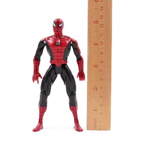 May include: A Spider-Man action figure, primarily red and black, stands upright against a white background. The figure is approximately 6 inches tall, with a wooden ruler placed beside it for scale. The figure's design is detailed, with a webbed pattern.