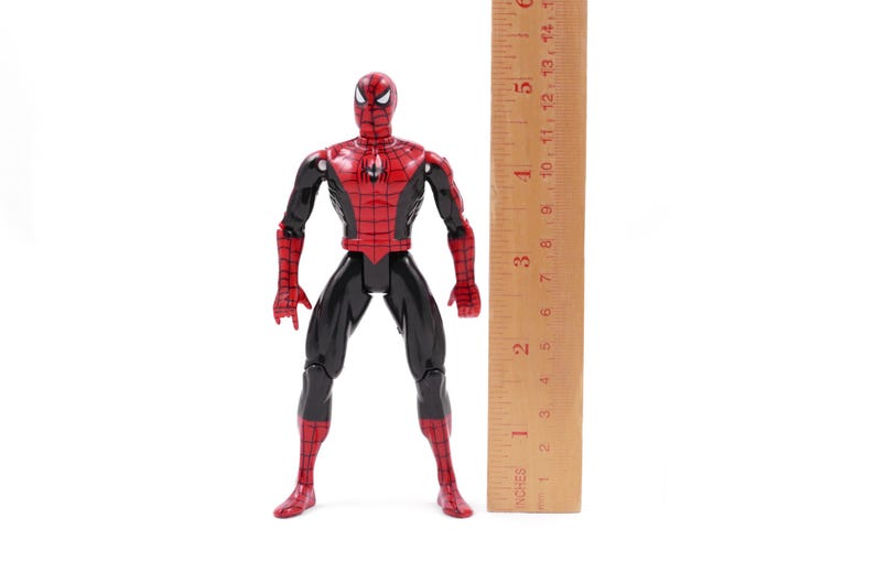 May include: A Spider-Man action figure, primarily red and black, stands upright against a white background. The figure is approximately 6 inches tall, with a wooden ruler placed beside it for scale. The figure's design is detailed, with a webbed pattern.