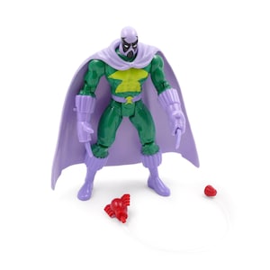 Puede incluir: Una figura de acci&oacute;n verde con capa, guantes y botas moradas. La figura tiene un emblema amarillo en el pecho y una m&aacute;scara morada. Un accesorio rojo est&aacute; en el suelo, conectado por una cuerda.