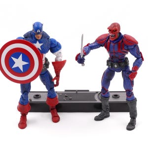 May include: Two action figures of Captain America and Red Skull. Captain America is in a blue and red costume, holding a shield. Red Skull is in a red and blue costume, holding a sword. Both figures are on a black base.