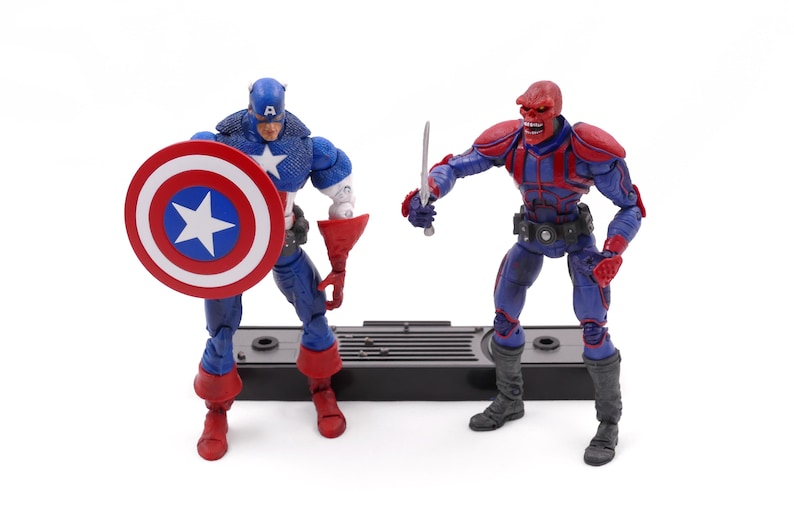 May include: Two action figures of Captain America and Red Skull. Captain America is in a blue and red costume, holding a shield. Red Skull is in a red and blue costume, holding a sword. Both figures are on a black base.