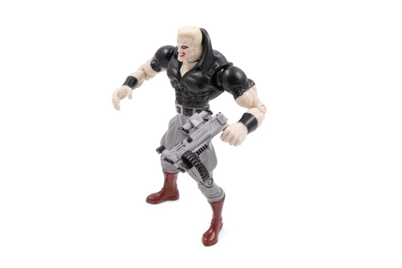 May include: Action figure of a muscular character with a pale complexion, wearing a black top, gray pants, and brown boots. The figure is holding a gray weapon and has black wristbands. The figure has a determined expression.