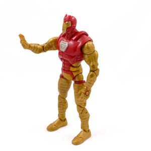 May include: A red and gold action figure of a superhero, posed with one arm extended. The figure has a red torso and head, with gold-colored arms and legs. The figure is standing on a white surface.