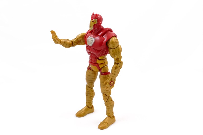 May include: A red and gold action figure of a superhero, posed with one arm extended. The figure has a red torso and head, with gold-colored arms and legs. The figure is standing on a white surface.