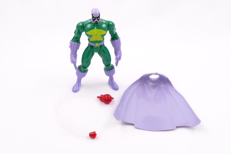 Puede incluir: Una figura de acci&oacute;n verde y amarilla con guantes, botas y m&aacute;scara moradas se encuentra junto a una capa de color lila claro y dos accesorios rojos. La figura tiene un dise&ntilde;o detallado y est&aacute; posada sobre un fondo blanco.