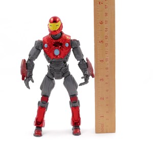 Op de afbeelding: Een rood en grijs actiefiguur van een superheld, rechtopstaand. Het figuur heeft een gele helm en is ongeveer 18 cm hoog. Een houten liniaal is naast het figuur geplaatst voor de schaal.