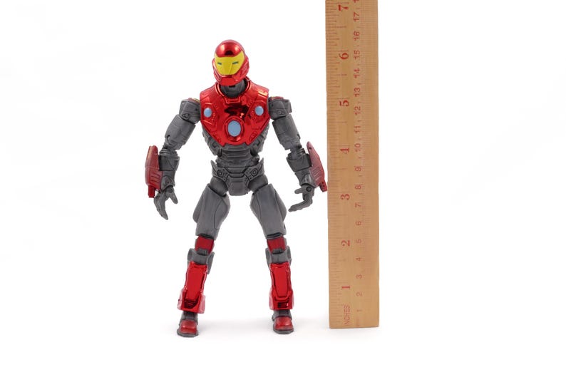 Op de afbeelding: Een rood en grijs actiefiguur van een superheld, rechtopstaand. Het figuur heeft een gele helm en is ongeveer 18 cm hoog. Een houten liniaal is naast het figuur geplaatst voor de schaal.