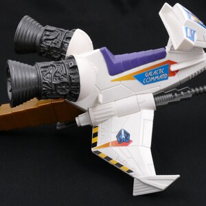 Op de afbeelding: Een wit en paars speelgoed ruimteschip met de tekst "GALACTIC COMMAND" aan de zijkant. Het speelgoed heeft twee grote grijze motoren en een bruine verlenging. De vleugels hebben zwart-gele strepen en een blauw embleem.