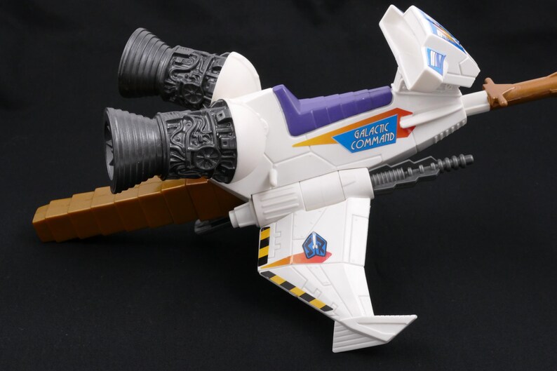 Op de afbeelding: Een wit en paars speelgoed ruimteschip met de tekst "GALACTIC COMMAND" aan de zijkant. Het speelgoed heeft twee grote grijze motoren en een bruine verlenging. De vleugels hebben zwart-gele strepen en een blauw embleem.
