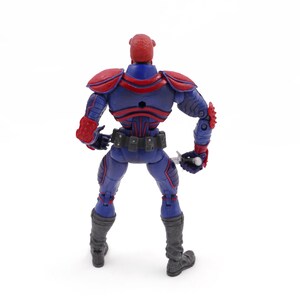 May include: A blue and red action figure with a helmet, shoulder pads, and gloves. The figure has a utility belt and gray boots. It is holding a silver weapon. The figure is standing on a white surface.