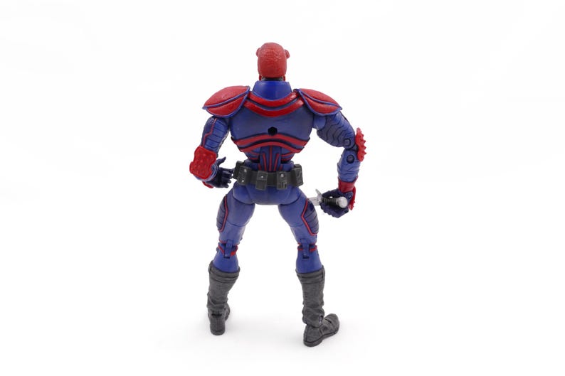 May include: A blue and red action figure with a helmet, shoulder pads, and gloves. The figure has a utility belt and gray boots. It is holding a silver weapon. The figure is standing on a white surface.