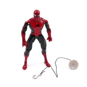 May include: A Spider-Man action figure in a standing pose, featuring a red and black costume. The figure has a web-slinging accessory with a hook and a disc-shaped component. The toy is set against a white background.
