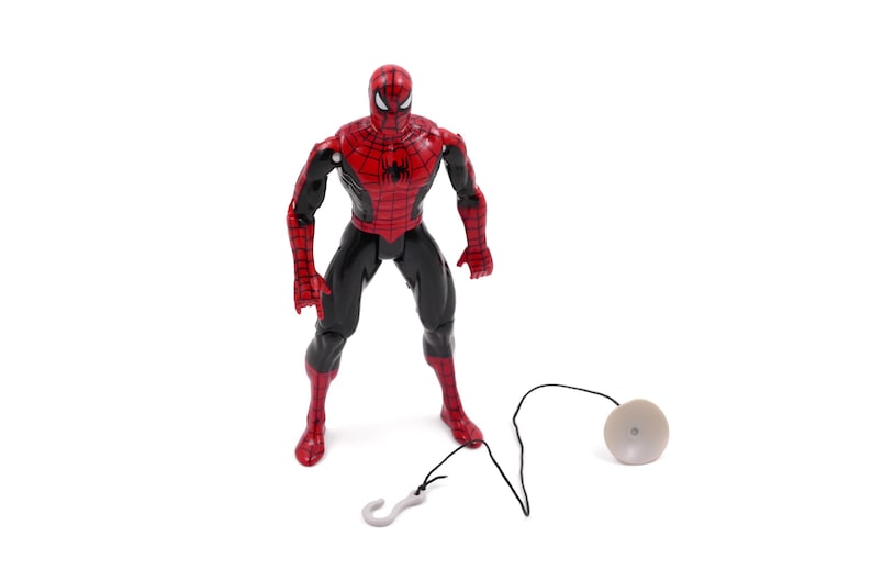 May include: A Spider-Man action figure in a standing pose, featuring a red and black costume. The figure has a web-slinging accessory with a hook and a disc-shaped component. The toy is set against a white background.