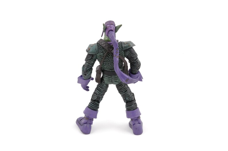 K&ouml;nnte beinhalten: Eine Green Goblin-Actionfigur, von hinten gesehen, steht auf einer wei&szlig;en Oberfl&auml;che. Die Figur ist &uuml;berwiegend gr&uuml;n und lila, mit detaillierter R&uuml;stung und einer langen, lila Kapuze. Die Figur hat eine detaillierte Textur.