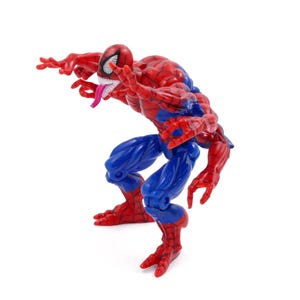 May include: A red, blue, and white action figure of a superhero with a white-eyed mask, a long pink tongue, and a muscular build. The figure is in a crouching pose, with its arms outstretched.