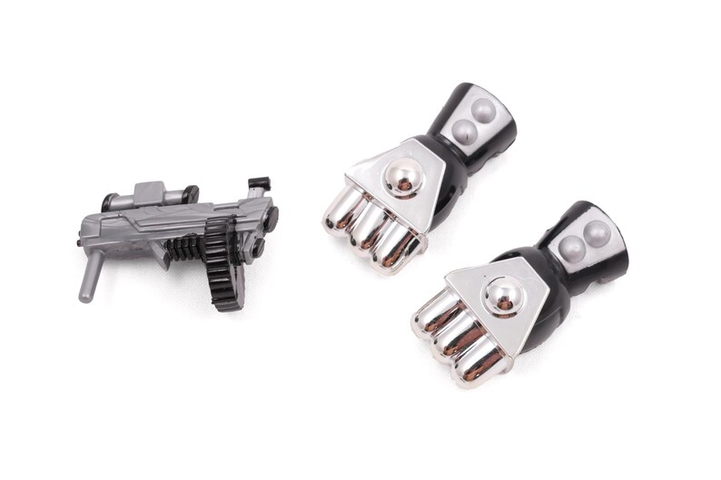 May include: A gray toy machine gun with a black ammunition belt and two black and silver robotic fists. The toy gun has a scope and a handle. The fists have silver knuckles and black cuffs.