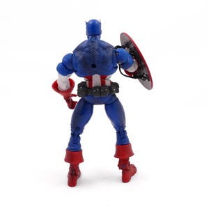 May include: A Captain America action figure, viewed from the back, stands on a white surface. The figure is primarily blue with red and white accents. It holds a red and silver shield and wears red boots. The figure is a collectible toy.