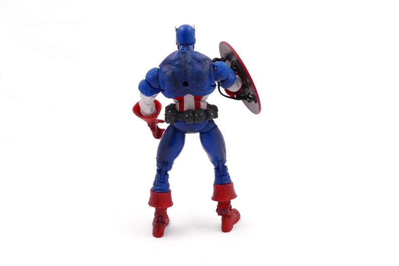May include: A Captain America action figure, viewed from the back, stands on a white surface. The figure is primarily blue with red and white accents. It holds a red and silver shield and wears red boots. The figure is a collectible toy.