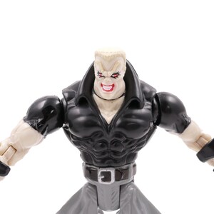 May include: A plastic action figure of a muscular character with a stern expression. The figure wears a black jacket, gray pants, and a silver belt. The figure's arms are outstretched, and it has a menacing grin.