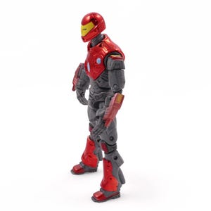 Op de afbeelding: Een rood en grijs actiefiguur van een superheld in een staande pose. De figuur heeft een rode helm met een geel vizier, rode bepantsering op de borst en armen, en grijze benen. De figuur staat op een witte achtergrond.