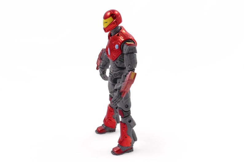 Op de afbeelding: Een rood en grijs actiefiguur van een superheld in een staande pose. De figuur heeft een rode helm met een geel vizier, rode bepantsering op de borst en armen, en grijze benen. De figuur staat op een witte achtergrond.