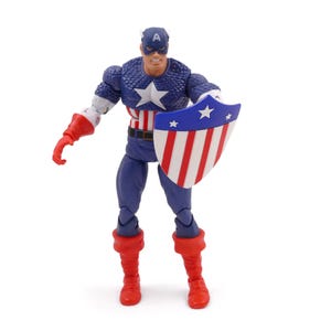 K&ouml;nnte beinhalten: Eine Captain America-Actionfigur vor wei&szlig;em Hintergrund. Die Figur tr&auml;gt ein blau-wei&szlig;es Kost&uuml;m mit einem Stern auf der Brust und einem passenden Helm. Sie h&auml;lt einen Schild mit roten und wei&szlig;en Streifen und blauen Sternen.