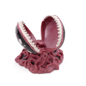 May include: A decorative, open-mouthed plant sculpture with a black exterior, white polka dots, and white teeth. The interior is a deep red color, and the base is a textured, reddish-brown. The sculpture is designed to resemble a carnivorous plant.