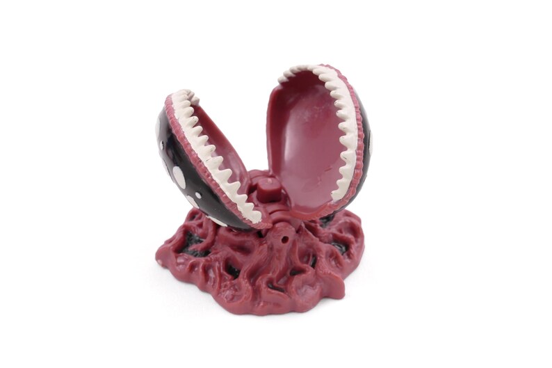 May include: A decorative, open-mouthed plant sculpture with a black exterior, white polka dots, and white teeth. The interior is a deep red color, and the base is a textured, reddish-brown. The sculpture is designed to resemble a carnivorous plant.