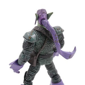 K&ouml;nnte beinhalten: Eine Green Goblin Actionfigur, von hinten gesehen, mit einer lila Kapuze und langen, flie&szlig;enden lila Haaren. Die Figur tr&auml;gt einen detaillierten gr&uuml;n-grauen Anzug mit einer kleinen braunen Tasche. Die Figur ist vor einem wei&szlig;en Hintergrund posiert.