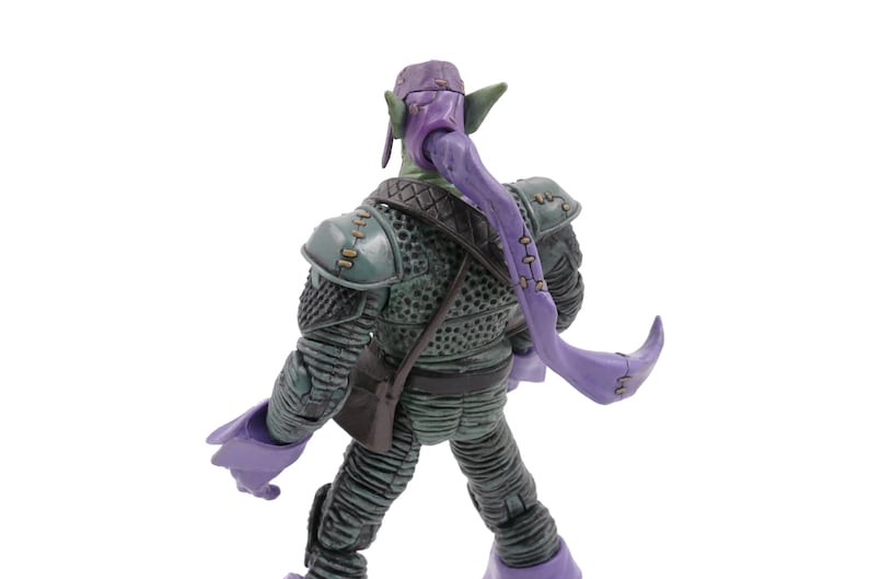 K&ouml;nnte beinhalten: Eine Green Goblin Actionfigur, von hinten gesehen, mit einer lila Kapuze und langen, flie&szlig;enden lila Haaren. Die Figur tr&auml;gt einen detaillierten gr&uuml;n-grauen Anzug mit einer kleinen braunen Tasche. Die Figur ist vor einem wei&szlig;en Hintergrund posiert.