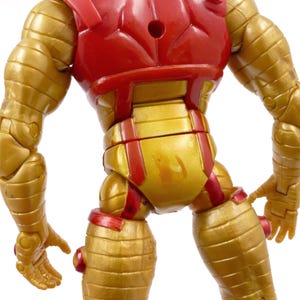 May include: A gold and red action figure of a superhero, viewed from the back. The figure has a gold metallic finish with red accents on the chest and joints. The figure's design features segmented armor and detailed musculature.