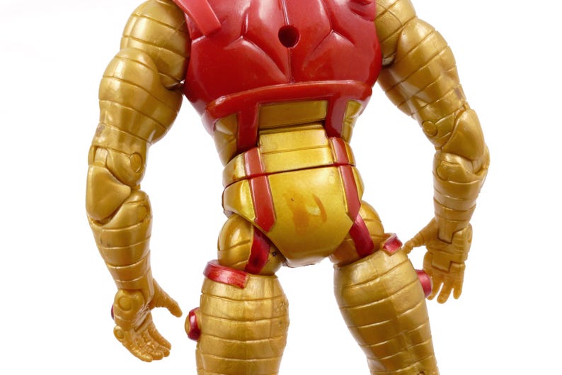 May include: A gold and red action figure of a superhero, viewed from the back. The figure has a gold metallic finish with red accents on the chest and joints. The figure's design features segmented armor and detailed musculature.