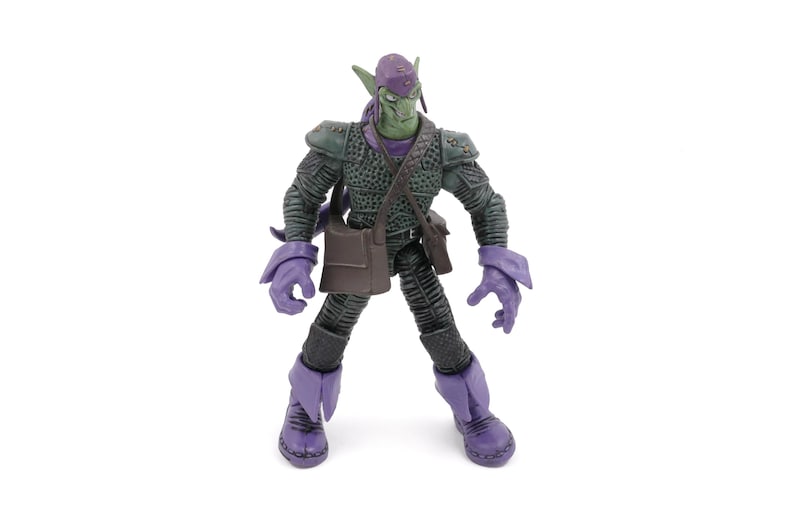 K&ouml;nnte beinhalten: Eine Green Goblin-Actionfigur mit lila Kapuze, Handschuhen und Stiefeln. Die Figur hat ein gr&uuml;nes Gesicht und ein dunkelgr&uuml;nes Outfit mit einer braunen Umh&auml;ngetasche. Die Figur steht auf einer wei&szlig;en Oberfl&auml;che.