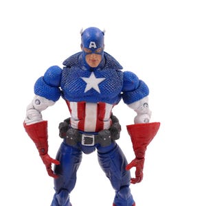 May include: A Captain America action figure in a heroic pose. The figure is predominantly blue, red, and white, with a star on the chest and a helmet. The figure has a detailed design and is set against a white background.