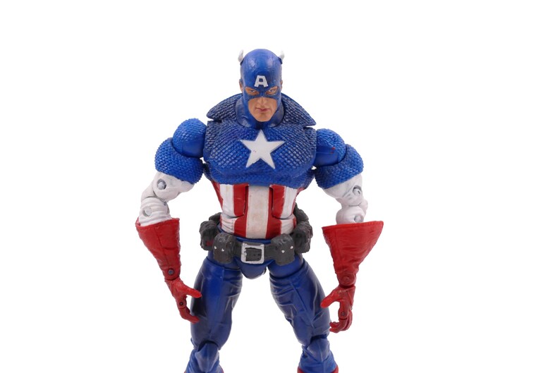 May include: A Captain America action figure in a heroic pose. The figure is predominantly blue, red, and white, with a star on the chest and a helmet. The figure has a detailed design and is set against a white background.