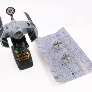 May include: A gray and black Star Wars TIE Interceptor toy with green lights on the bottom. The toy is on a black base with the words "Star Wars" in gold lettering. The toy is next to a gray and white paper model of a Star Destroyer.